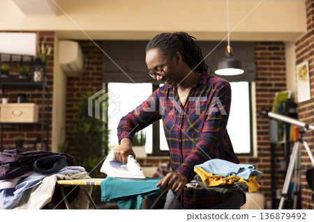 Smiling black housewife using electric iron to press clean shirt, removing wrinkles after laundry. Cheerful and focused, african american woman completes household chores in modern apartment. Smiling black housewife using electric iron to press clean shirt, removing wrinkles after laundry. Cheerful and focused, african american woman completes household chores in modern apartment. 136879492