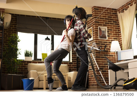 Beautiful young couple cleaning living room together, turning chores into fun while singing and dancing in brick wall apartment. Carefree boyfriend and girlfriend enjoying housework at home. 136879500