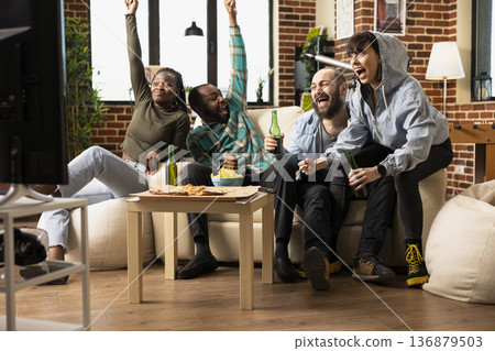 Multiethnic men and women cheer on couch over victory of favorite sports team. Weekend tv entertainment brings joy, bonding and celebration during relaxed home hangout with friends. 136879503