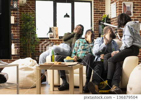 African american and caucasian friends sit on sofa with beer, reacting with fear to frightening scene on television. Multiethnic peers look away from TV, bonding during horror movie hangout at home. African american and caucasian friends sit on sofa with beer, reacting with fear to frightening scene on television. Multiethnic peers look away from TV, bonding during horror movie hangout at home. 136879504