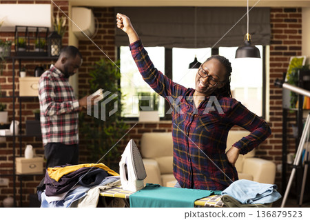 Black woman irons clothes and sings in brick wall apartment. Beautiful housewife joyfully dances with one hand in air, taking short break from ironing pile of shirts while multitasking during 136879523