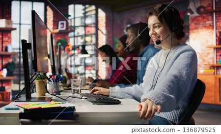 Friendly call center employee talking in audio headset, assisting customers in office. Happy customer support operator communicating with clients during telephone call, camera B close up 136879525