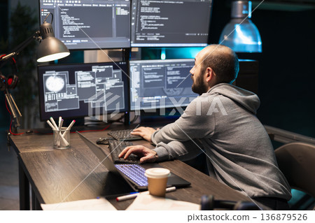 Male developer leans toward several monitors while reviewing complex lines of code during late night work session. Concentrated software engineer works in tech office, refining tasks with precision. 136879526