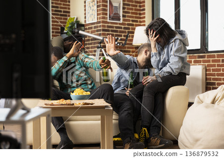 Diverse friends sit on couch covering faces as scary scene plays on television. African american and caucasian friends not looking at TV screen during horror movie hangout at home. Diverse friends sit on couch covering faces as scary scene plays on television. African american and caucasian friends not looking at TV screen during horror movie hangout at home. 136879532