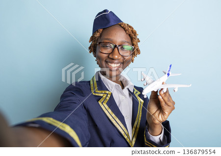 Black female air hostess playfully poses for selfie while holding model aircraft. Cheerful cabin crew member enjoys documenting experience in aviation industry in blue background studio. 136879554