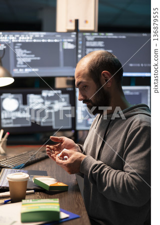 Male IT worker holds medication at desk, managing fatigue after long hours solving technical problems. Caucasian programmer checks pills while surrounded by monitors displaying complex system code. Male IT worker holds medication at desk, managing fatigue after long hours solving technical problems. Caucasian programmer checks pills while surrounded by monitors displaying complex system code. 136879555