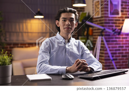 Confident professional man and entrepreneur sits at desk with computer keyboard, looking at camera. Portrait of focused asian male executive and leader working remotely in modern apartment. Confident professional man and entrepreneur sits at desk with computer keyboard, looking at camera. Portrait of focused asian male executive and leader working remotely in modern apartment. 136879557