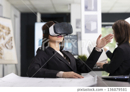 Professional female architect sits in office wearing VR headset and examining floor plans for building project. Woman examining 3D layouts and planning workflow, using digital and immersive tools. Professional female architect sits in office wearing VR headset and examining floor plans for building project. Woman examining 3D layouts and planning workflow, using digital and immersive tools. 136879581
