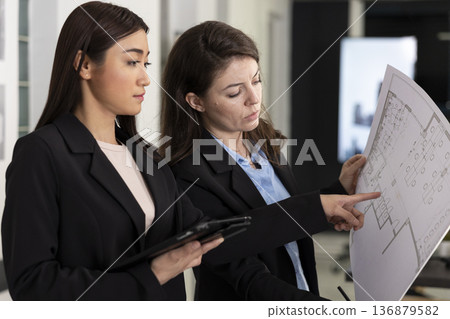 Diverse coworkers analyze architectural drawings, looking at floor plan details and structural elements. Asian and caucasian architects reviewing building measurements and taking notes on tablet. Diverse coworkers analyze architectural drawings, looking at floor plan details and structural elements. Asian and caucasian architects reviewing building measurements and taking notes on tablet. 136879582