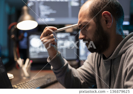 Exhausted tech freelancer leans forward holding pen to forehead while reviewing coding errors. Sad male programmer feeling fatigued and stressed after hours of intense programming work. 136879625