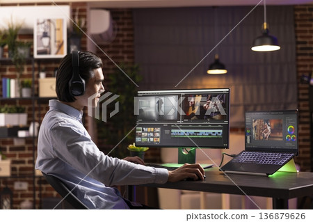 Young man focuses on matching audio with video footage during post production. Asian male filmmaker enjoys music during editing process on digital devices, working from cozy home office studio. Young man focuses on matching audio with video footage during post production. Asian male filmmaker enjoys music during editing process on digital devices, working from cozy home office studio. 136879626