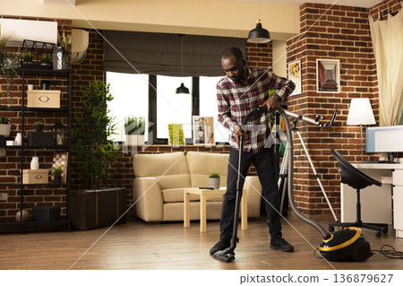 African American male homeowner vacuuming living room floor in bright apartment. Young black man holding household appliance, focused on keeping brick wall home clean and tidy during weekend chores. African American male homeowner vacuuming living room floor in bright apartment. Young black man holding household appliance, focused on keeping brick wall home clean and tidy during weekend chores. 136879627