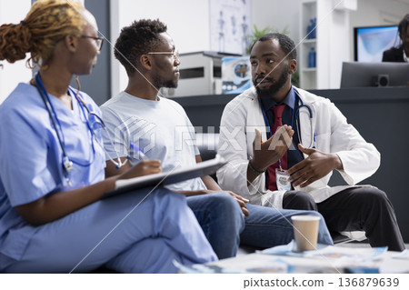 Black male doctor in lab coat speaks with patient during clinic appointment, reviewing documents and discussing health concerns. Female nurse takes notes as physician explains treatment information. 136879639