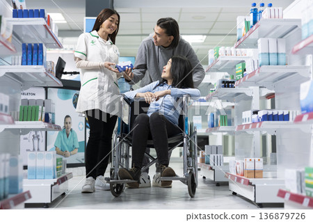 Female pharmacist provides inclusive healthcare service to woman in wheelchair and her companion. Focus on pharmacy accessibility, patient support and professional medical guidance in store. Female pharmacist provides inclusive healthcare service to woman in wheelchair and her companion. Focus on pharmacy accessibility, patient support and professional medical guidance in store. 136879726