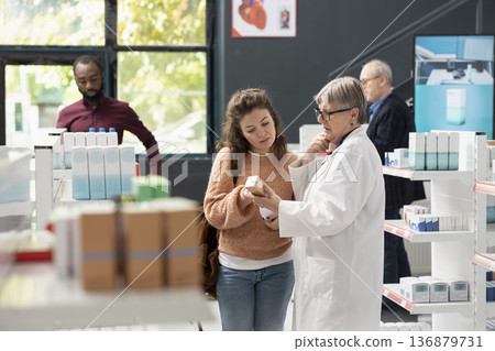 Female pharmacist explains proper medication usage to female customer holding pharmaceutical product. Medical professional offers guidance on supplements and prescriptions in local pharmacy. Female pharmacist explains proper medication usage to female customer holding pharmaceutical product. Medical professional offers guidance on supplements and prescriptions in local pharmacy. 136879731