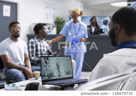 Male physician sits in clinic lobby, conducting remote video consultation with caucasian colleague. Diverse male doctors collaborate online to review medical tests and coordinate clinical procedures. 136879735