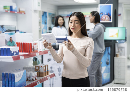 Female customer carefully compares over the counter medicine while standing in drugstore aisle. Adult woman reads product labels and information to ensure she selects correct medication for her needs. Female customer carefully compares over the counter medicine while standing in drugstore aisle. Adult woman reads product labels and information to ensure she selects correct medication for her needs. 136879738