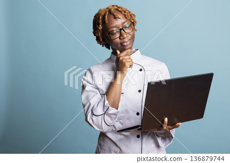 Professional cook with laptop, concentrates on digital research and plans gourmet dishes. African American chef stands with thoughtful expression, browsing online ingredients and meal inspirations. Professional cook with laptop, concentrates on digital research and plans gourmet dishes. African American chef stands with thoughtful expression, browsing online ingredients and meal inspirations. 136879744