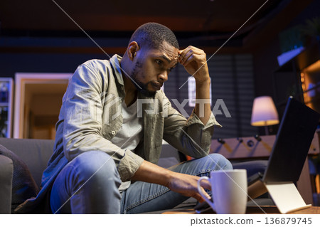 African american adult receiving some bad news while doing remote work tasks, feeling overworked and under pressure to complete all his assignments. Young person dealing with burnout. African american adult receiving some bad news while doing remote work tasks, feeling overworked and under pressure to complete all his assignments. Young person dealing with burnout. 136879745