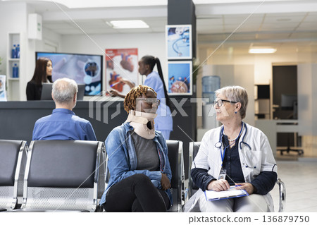 Attentive doctor providing friendly assistance to patient with neck brace seated in hospital waiting room. Retired female physician wearing lab coat reassuring injured black woman of speedy recovery. Attentive doctor providing friendly assistance to patient with neck brace seated in hospital waiting room. Retired female physician wearing lab coat reassuring injured black woman of speedy recovery. 136879750