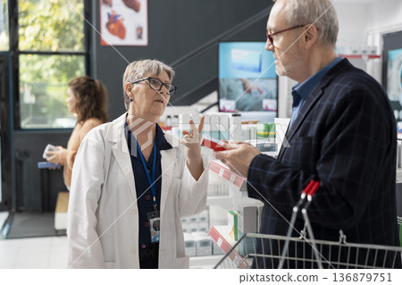 Senior pharmacist advises male customer on taking medication according to prescription. Female drugstore attendant guides man holding basket in choosing beneficial health supplements and products. Senior pharmacist advises male customer on taking medication according to prescription. Female drugstore attendant guides man holding basket in choosing beneficial health supplements and products. 136879751