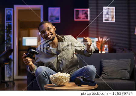 African american successful guy enjoys the gaming victory at home, celebrating and feeling confident about his success. Cheerful joyous black man enjoying free time and a gaming win. African american successful guy enjoys the gaming victory at home, celebrating and feeling confident about his success. Cheerful joyous black man enjoying free time and a gaming win. 136879754