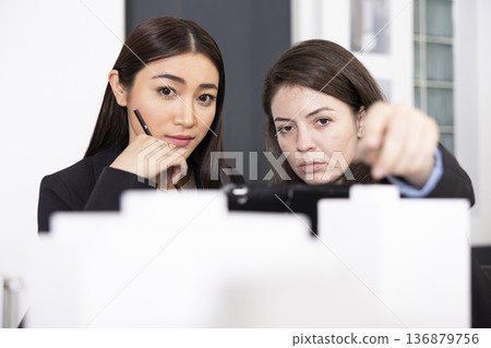Young women in focus on developing architectural layouts and drafting floor plan ideas. Caucasian architect explains 3d building model to coworker in preparation of construction briefing with client. Young women in focus on developing architectural layouts and drafting floor plan ideas. Caucasian architect explains 3d building model to coworker in preparation of construction briefing with client. 136879756