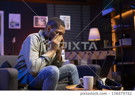 Young black person overworked with a headache in living room, working remotely under stress and pressure from his boss. Young adult frustrated and worried, struggling alone with tasks. 136879782