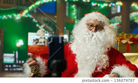Santa Claus using smartphone on tripod to film vlog opening letters from kids. Older person wearing Christmas costume recording video with phone selfie camera, reading letters from children, camera A Santa Claus using smartphone on tripod to film vlog opening letters from kids. Older person wearing Christmas costume recording video with phone selfie camera, reading letters from children, camera A 136879787