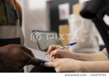 Close up of woman reviewing inventory list before packing goods for delivery, confirming the order processing operation sin house and managing the logistics side of the small business depot. Close up of woman reviewing inventory list before packing goods for delivery, confirming the order processing operation sin house and managing the logistics side of the small business depot. 136879791