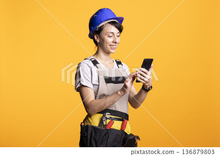Caucasian female builder in safety helmet cheerfully using cellphone, reading funny message or instruction. Woman contractor stands with tool belt, reflecting onsite coordination and mobile updates. Caucasian female builder in safety helmet cheerfully using cellphone, reading funny message or instruction. Woman contractor stands with tool belt, reflecting onsite coordination and mobile updates. 136879803