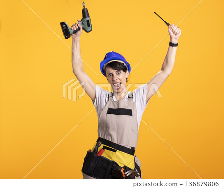 Happy white woman in overalls and tool belt lifts drill gun and screwdriver with excitement. Caucasian female builder dances and sings in studio, having fun with construction tools. Happy white woman in overalls and tool belt lifts drill gun and screwdriver with excitement. Caucasian female builder dances and sings in studio, having fun with construction tools. 136879806