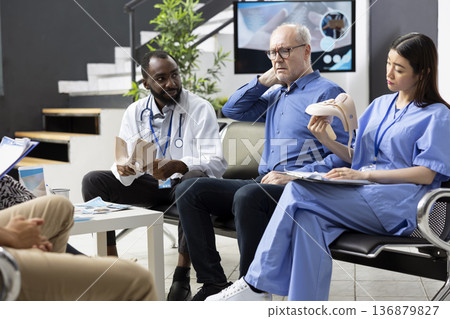 Caucasian man points to neck and shoulder pain during consultation with medical specialists, helping guide diagnosis and treatment for upper body discomfort. Diverse physicians speaking with patient. Caucasian man points to neck and shoulder pain during consultation with medical specialists, helping guide diagnosis and treatment for upper body discomfort. Diverse physicians speaking with patient. 136879827
