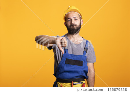 Male builder gestures thumbs down, expressing dissatisfaction or disapproval. Portrait of construction worker on yellow background representing evaluation, feedback or negative review at building site Male builder gestures thumbs down, expressing dissatisfaction or disapproval. Portrait of construction worker on yellow background representing evaluation, feedback or negative review at building site 136879838