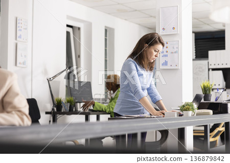 Corporate female professional looks at laptop, reviewing project details and planning upcoming work tasks. Caucasian woman manager standing and checking business information on digital device. Corporate female professional looks at laptop, reviewing project details and planning upcoming work tasks. Caucasian woman manager standing and checking business information on digital device. 136879842