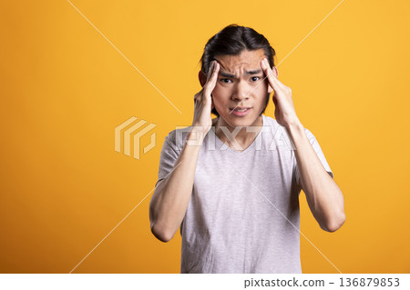 Frustrated asian man massaging temples while feeling overwhelmed against isolated background. Young adult male expresses stress or tension, representing anxiety, headache burnout or overthinking. Frustrated asian man massaging temples while feeling overwhelmed against isolated background. Young adult male expresses stress or tension, representing anxiety, headache burnout or overthinking. 136879853