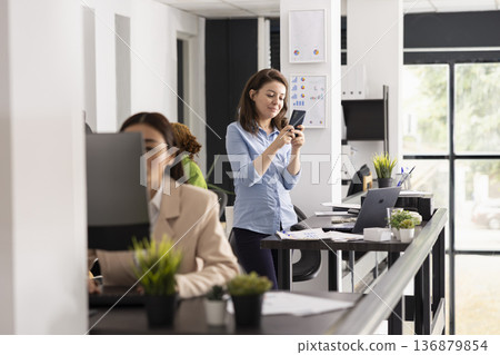 Smiling businesswoman stands in office, taking short break and browsing social media on smartphone. Female employee multitasks between texting colleagues and reviewing project information on devices. Smiling businesswoman stands in office, taking short break and browsing social media on smartphone. Female employee multitasks between texting colleagues and reviewing project information on devices. 136879854
