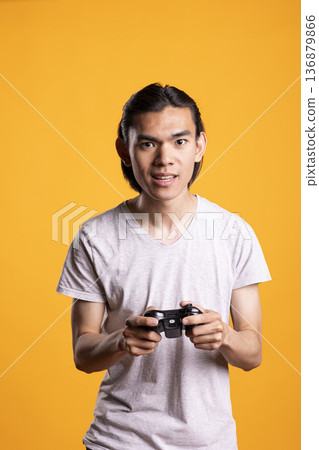 Angry man clenches game controller tense facial expression, reacting to gaming defeat. Unhappy player shows irritation and focus while participating in intense online multiplayer session. 136879866