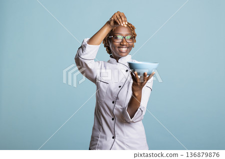 African american professional cook happily sprinkling pinch of salt into bowl to enhance flavor. Happy female chef adds final touches to freshly prepared meal, showing joy and passion for cooking. African american professional cook happily sprinkling pinch of salt into bowl to enhance flavor. Happy female chef adds final touches to freshly prepared meal, showing joy and passion for cooking. 136879876