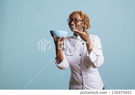 Female culinary expert posing against blue background, gently holding bowl of freshly prepared meal. Confident chef with eyes closed and satisfied smile, appreciating aroma of cooked food. Female culinary expert posing against blue background, gently holding bowl of freshly prepared meal. Confident chef with eyes closed and satisfied smile, appreciating aroma of cooked food. 136879881