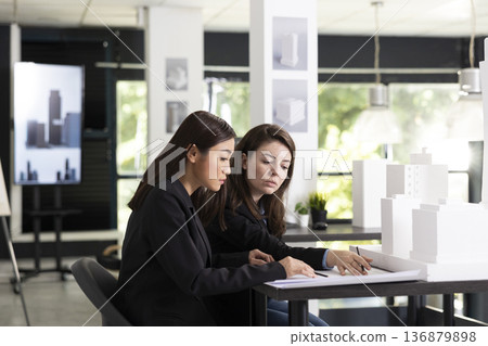 Female architects review blueprint details and discuss layout ideas in modern design studio. Dedicated diverse professionals examine architectural models, assessing measurements and design accuracy. Female architects review blueprint details and discuss layout ideas in modern design studio. Dedicated diverse professionals examine architectural models, assessing measurements and design accuracy. 136879898