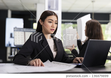 Asian architect concentrates on laptop, preparing reports and coordinating timelines for construction site evaluations. Professional woman brainstorms building ideas and researching online floor plans 136879899