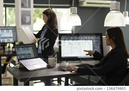 Young architectural designer analyzing building layout accuracy and technical drawing details on computer. Asian woman architect evaluating client requirements and preparing project reports in office. 136879901