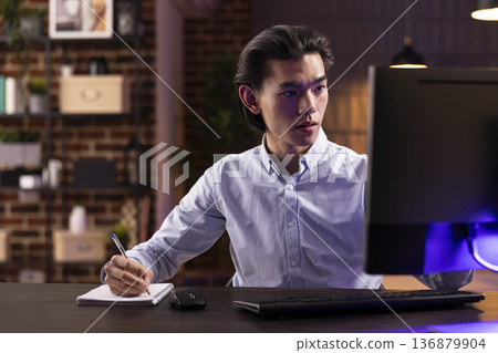 Focused male individual sits at table with computer and notebook, concentrating on freelance work. Handsome entrepreneur reviews marketing plans and improves business strategies online. Focused male individual sits at table with computer and notebook, concentrating on freelance work. Handsome entrepreneur reviews marketing plans and improves business strategies online. 136879904