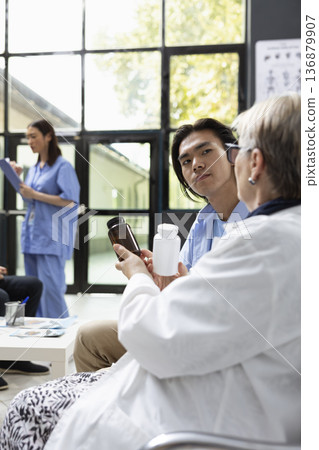Asian man consults physician in clinic lobby about prescribed medication. General practitioner explains dosage and side effects, emphasizing clear communication and professional healthcare guidance. 136879907