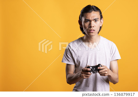 Furious man gripping gamepad tightly after losing online match. Unhappy male gamer reacting with frustration during tense gaming session, showing emotion and focus in modern studio. Furious man gripping gamepad tightly after losing online match. Unhappy male gamer reacting with frustration during tense gaming session, showing emotion and focus in modern studio. 136879917