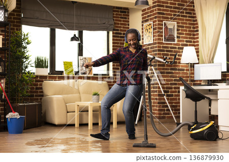 Positive housewife dances in living room, cleaning floor with vacuum and maintaining cleanliness in brick wall apartment. Happy young woman wears headphones, listening to music and vacuuming at home. Positive housewife dances in living room, cleaning floor with vacuum and maintaining cleanliness in brick wall apartment. Happy young woman wears headphones, listening to music and vacuuming at home. 136879930