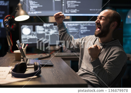 Joyful bearded programmer raising fists in celebration after solving debugging issue in tech workspace. Excited male software developer achieving coding breakthrough during night shift. 136879978