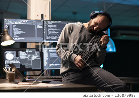Male programmer lifts computer keyboard like guitar while enjoying playful break in tech workspace. The lighthearted moment shows relaxed side of office culture amid demanding coding tasks. Male programmer lifts computer keyboard like guitar while enjoying playful break in tech workspace. The lighthearted moment shows relaxed side of office culture amid demanding coding tasks. 136879982