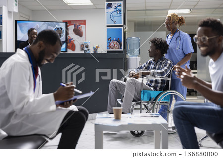 Nurse supports injured male patient seated in wheelchair while waiting for medical evaluation in hospital lobby. Doctor prepares documents and interacts with male visitor nearby in private clinic. 136880000